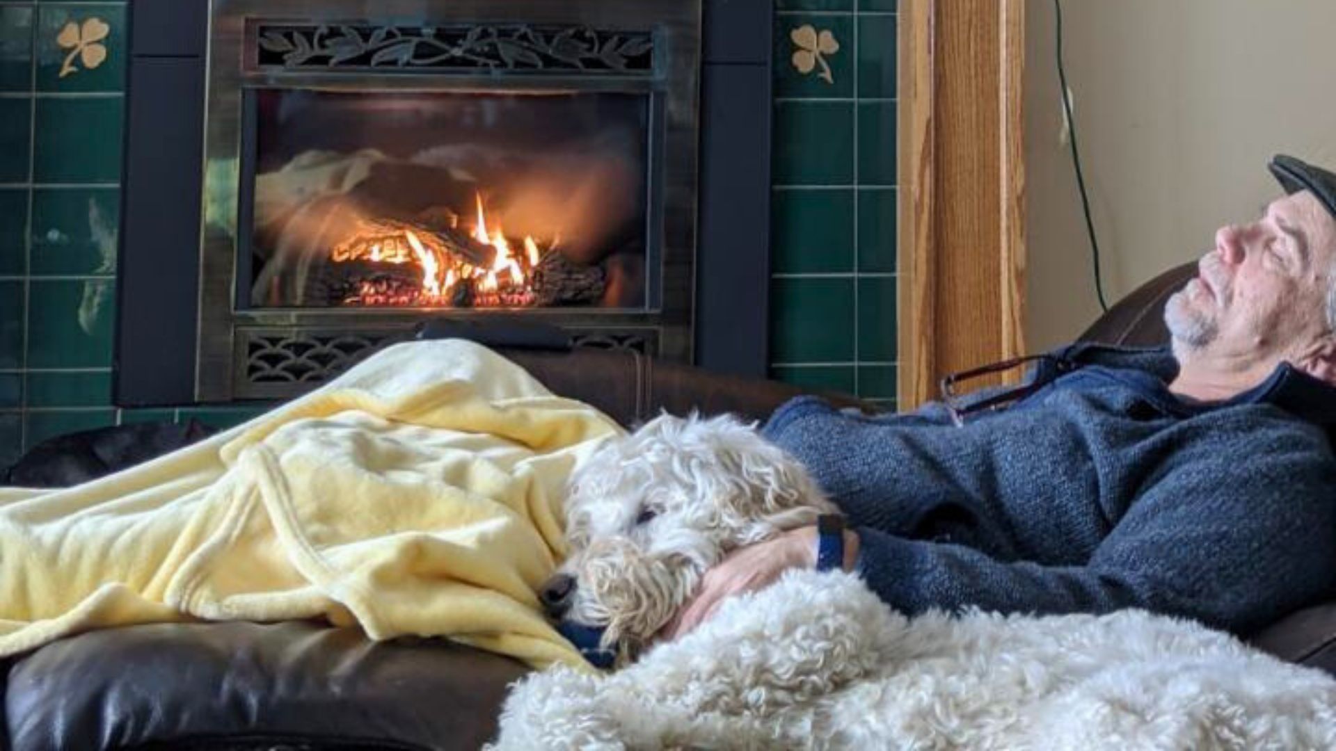 A man taking a nap on a leather chair with his white dog next to him, by a fireplace