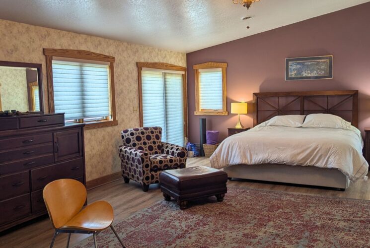 Spacious bedroom with king bed, area rug, sitting chair with ottoman, large dresser and windows with blinds