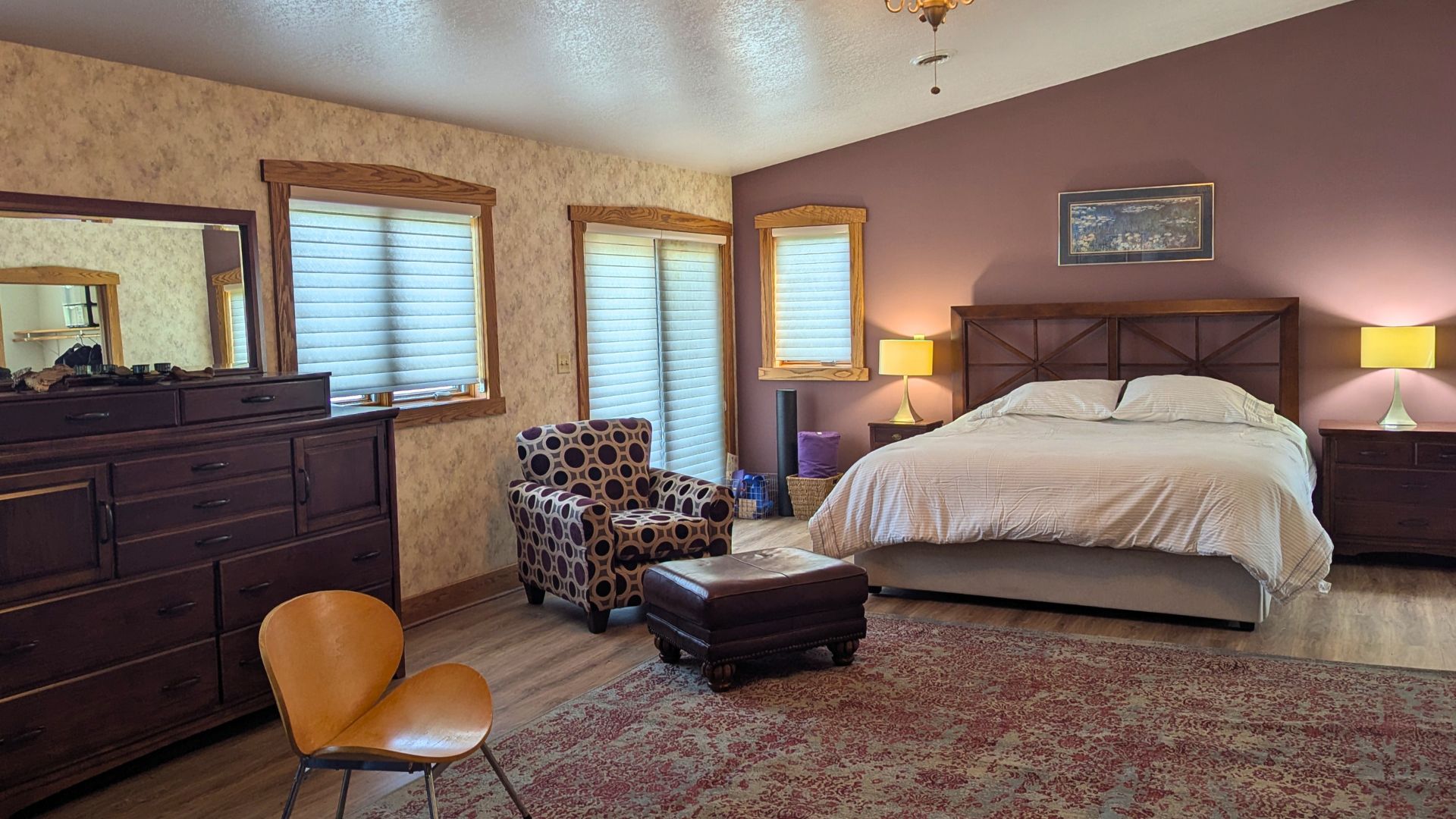 Spacious bedroom with king bed, area rug, sitting chair with ottoman, large dresser and windows with blinds