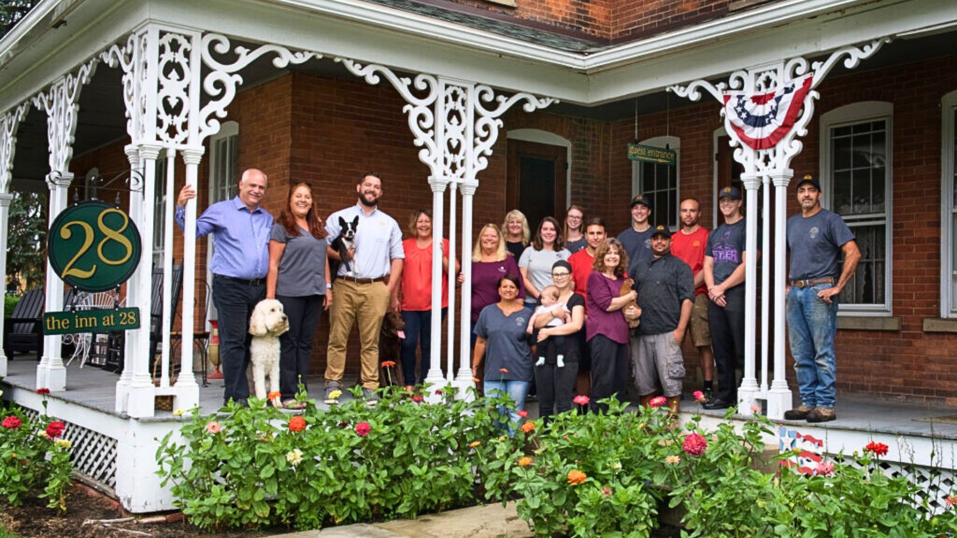 A large group of people standing on the outdoor wrap-around porch of a brick home with decorative white columns