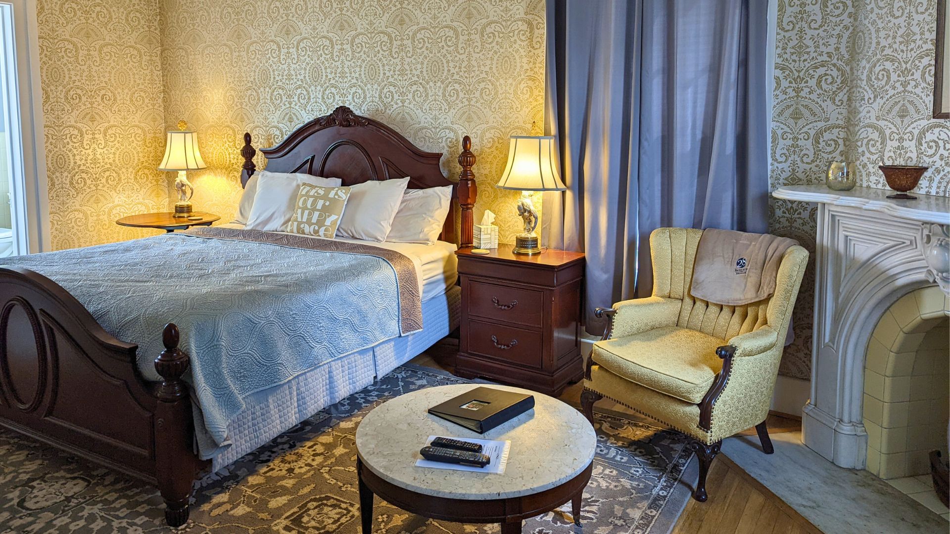 Elegant bedroom with bed with dark wood headboard and footboard, sitting chair and table by fireplace and decorative wallpaper