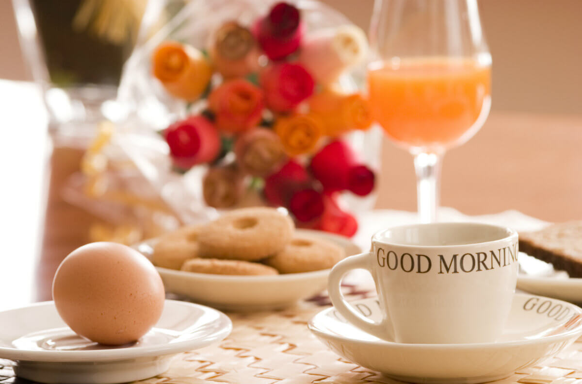 Breakfast table set with white cup, a whole, brown egg, donuts, glass of orange juice plus red and orange flowers