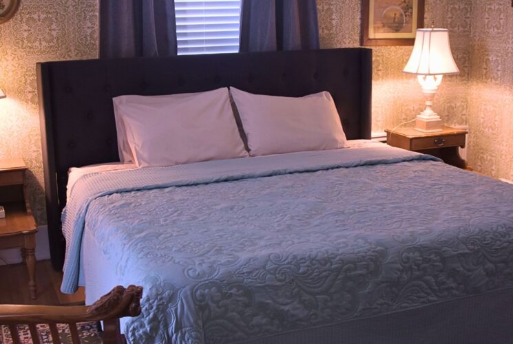 Bedroom with queen bed, tufted headboard, blue quilt, side tables with lamps, decorative wallpaper