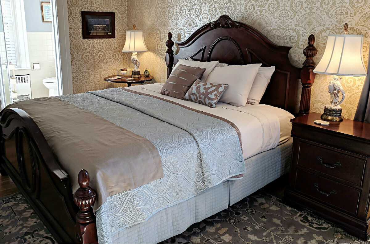 Bedroom with beautiful gold and white wallpaper, deep brown bed and night stands with white bathroom in background