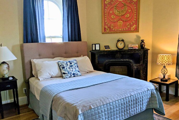 Elegant bedroom with queen bed, blue quilt, windows with blue curtains and decorative fireplace