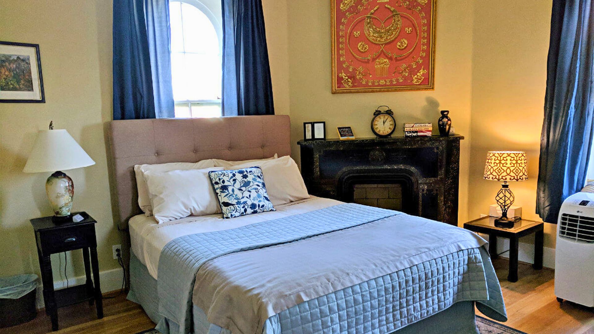 Elegant bedroom with queen bed, blue quilt, windows with blue curtains and decorative fireplace