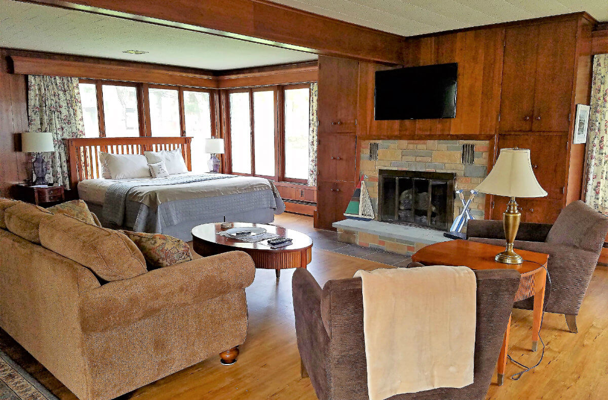 Large room with brown paneled walls with a tan couch, two brown chairs, a bed and stone fireplace