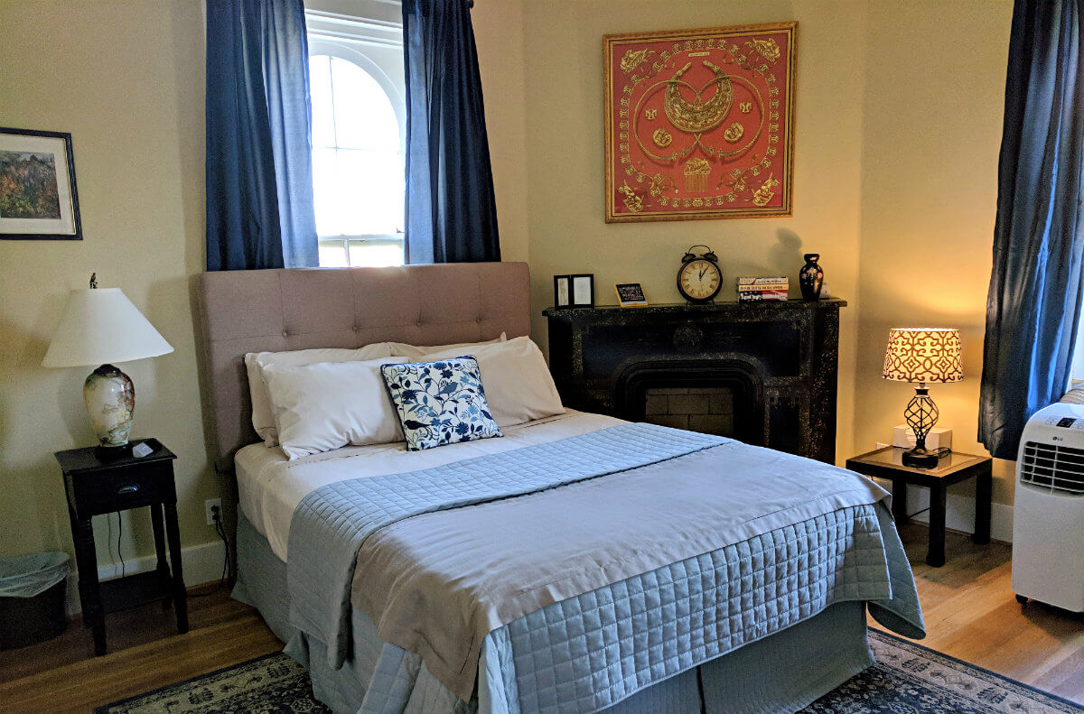 Bedroom with tan walls, dark blue curtains, blue patterned rug, and black fireplace with red wall hanging over mantle