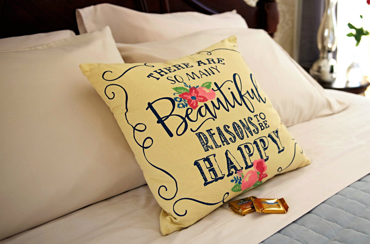 Four tan pillows behind cream pillow with blue words, there are so many beautiful reasons to be happy