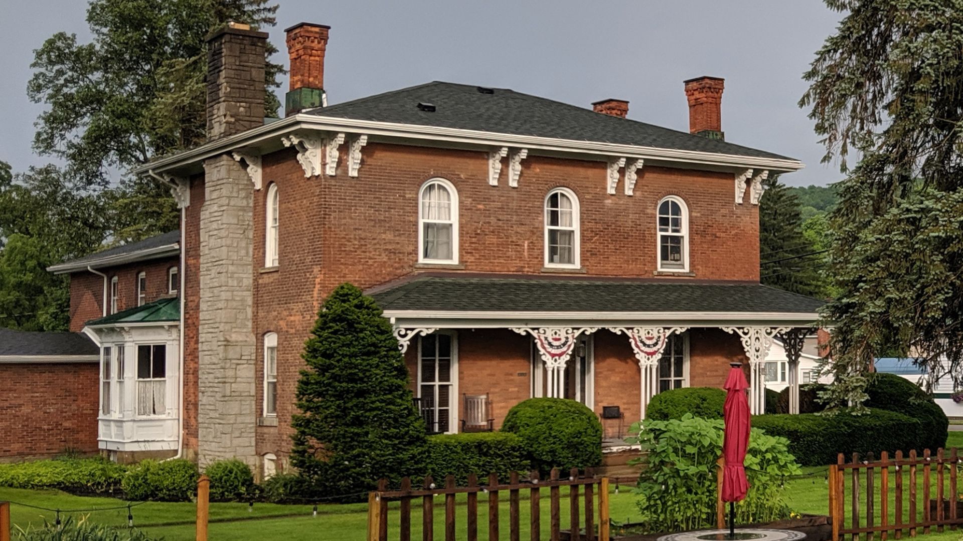 Large brown brick home with white corbels, green trees and bushes and arched windows