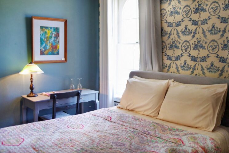 Bedroom with queen bed, quilt, decorative wallpaper, desk with chair by large window with curtains