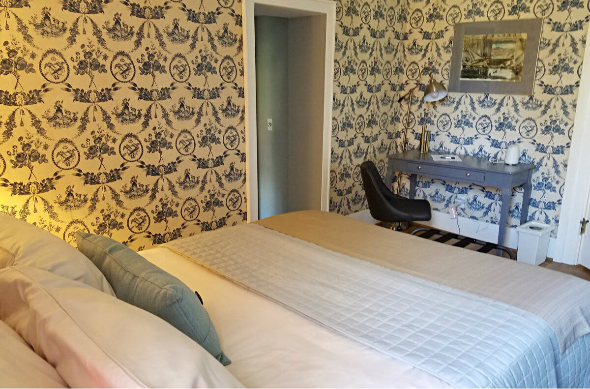 Bedroom with blue and white wallpaper, nightstand with bright light and a black luggage holder at the end of the bed