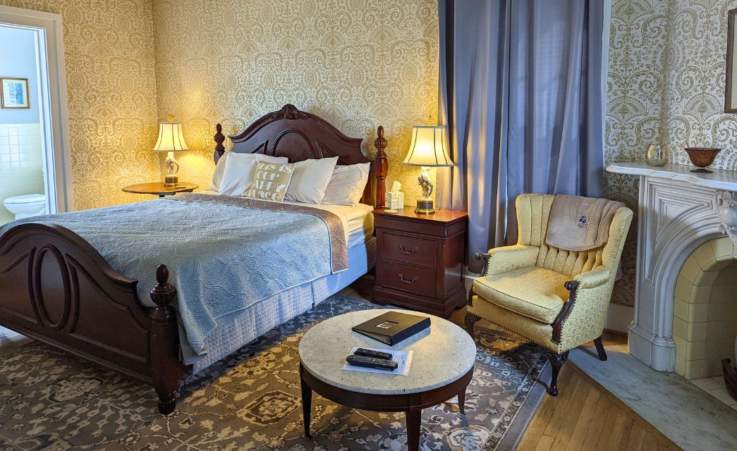 Elegant bedroom with king bed, side tables with lamps, wingback chair and round table by fireplace, decorative wallpaper and attached bathroom