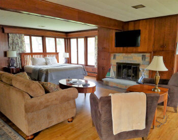 Large room with brown paneled walls with a tan couch, two brown chairs, a bed and stone fireplace