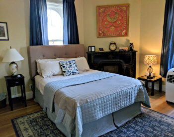 Bedroom with tan walls, dark blue curtains, blue patterned rug, and black fireplace with red wall hanging over mantle