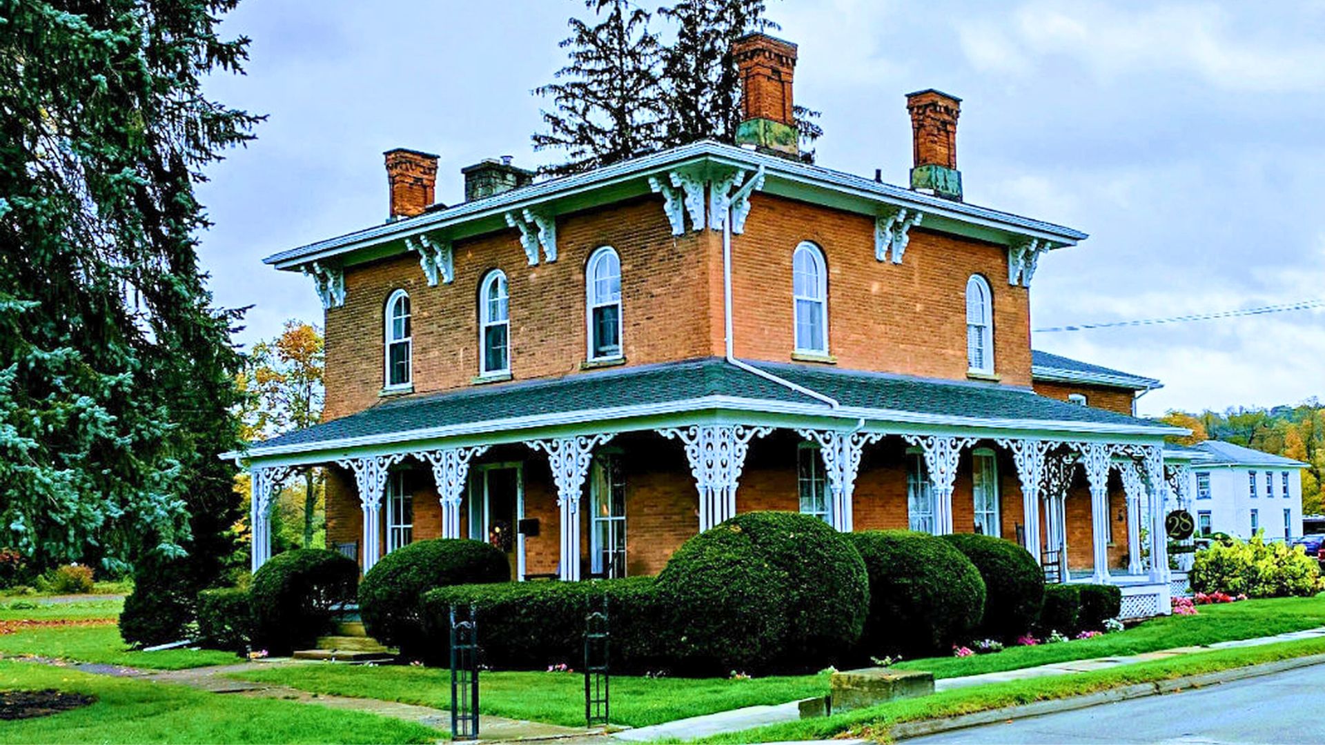 Exterior of a two-story brick home with wrap around porch, arched windows, decorative white columns, lush bushes and tall trees