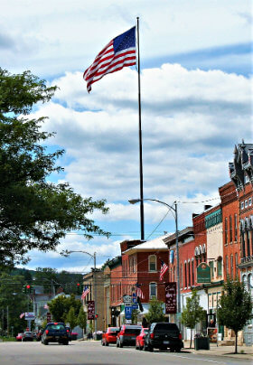 Main village street with several buildings, green trees and very tall flag pole with red, white and blue American flag