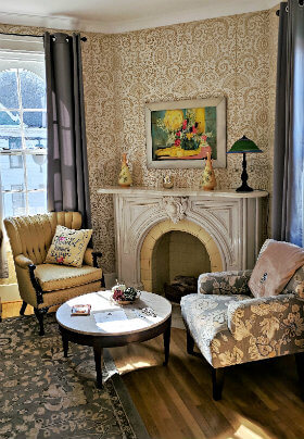 Seating area with ornate fireplace, gold patterned wallpaper and 2 chairs, one gold and one with grey design