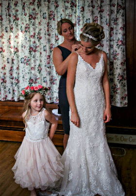 Woman in blue dress fastening white dress of a bride and a little girl standing beside them