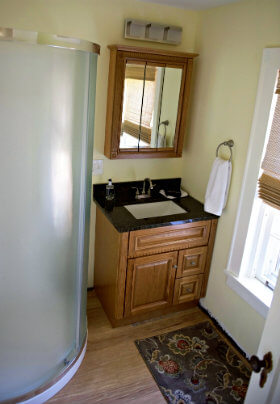 Bathroom with yellow walls, curved corner shower, patterned bathmat, mirror and brown vanity with black top