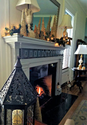 Black lantern decoration in foreground of white fireplace with blue tiles and gold, tree decorations on the mantle