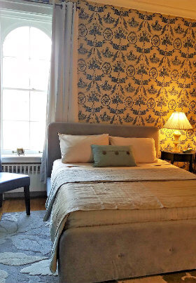 Bedroom with large, white, arched window, blue and tan bed and blue and white, patterned wallpaper