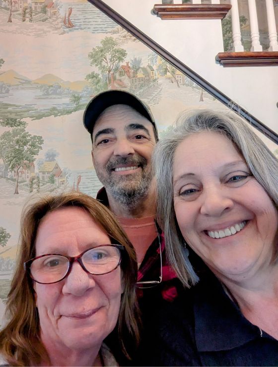 Two women and one man gathered for a picture by a wall with decorative wallpaper